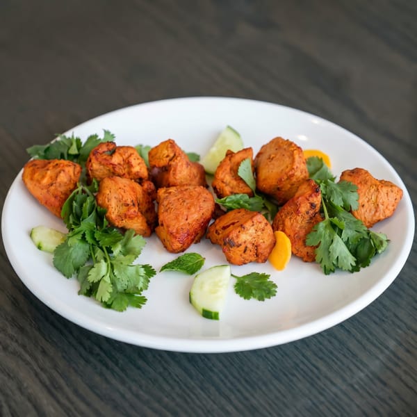 Orange chicken tikka pieces garnished with cilantro, mint, cucumber, and bell pepper on a white plate.
