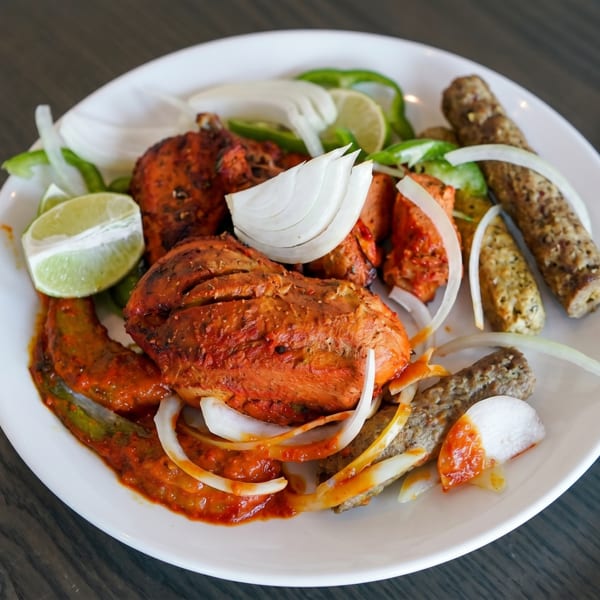 A white plate holds tandoori chicken, seekh kebabs, onions, green peppers, and lime wedges.