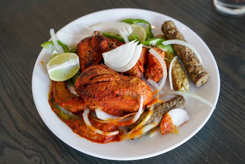 A white plate holds tandoori chicken, seekh kebabs, onions, green peppers, and lime wedges.
