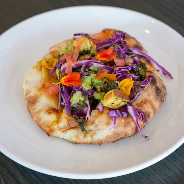 Flatbread topped with purple cabbage, green vegetables, and edible orange and yellow flowers on a white plate.